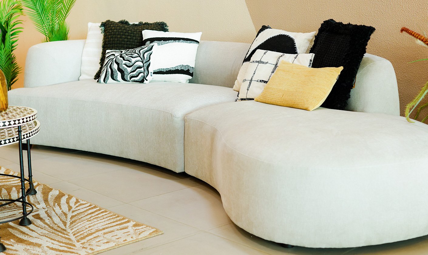 Modern curved beige sofa with patterned black-and-white cushions and a yellow accent pillow, styled with indoor plants and nested side tables.